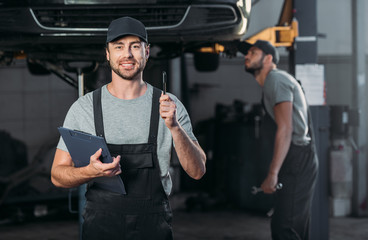 smiling mechanic wirh clipboard having idea, while colleague working in workshop behind