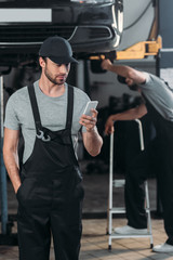 professional auto mechanic using smartphone, while colleague working in workshop behind