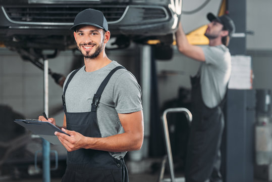 smiling mechanic writing in clipboard, while colleague working in workshop behind