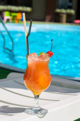 glass with an orange drink on the edge of a blue pool.