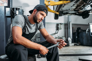 tired mechanic holding wrench and sitting in auto repair shop