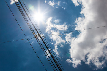 Fototapeta premium Electric and telecommunication wires with bright sun in a blue sky with clouds on the background. No people.