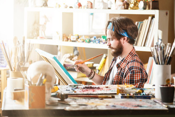 Young caucasian male artist in blue bandana drawing with brush and oil on canvas in his sunny studio