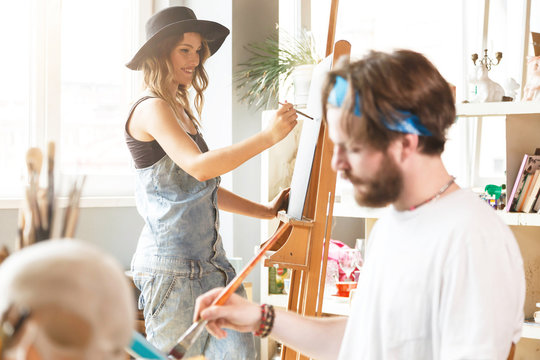 Caucasian dark-haired hipster male artist in blue bandana sitting and drawing with brush on canvas in his light modern studio, his colleague drawing behind the easel near the window