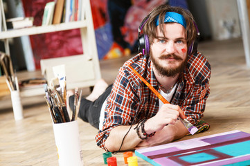 Handsome dark-haired bearded hipster male artist in headphones and blue bandana lies on the floor...