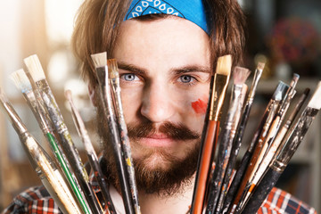 Cheerful dark-haired caucasian bearded hipster male artist in blue bandana hoilding many brushes in...