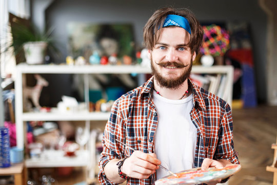 Caucasian dark-haired bearded hipster male artist in blue bandana mixed colors on the palette in his contemporary studio with many paintings