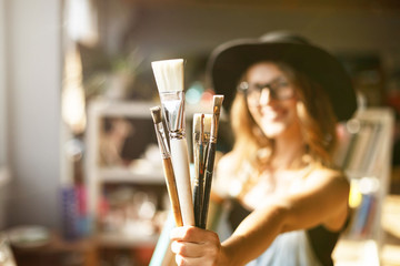 Cute caucasian long-hair female artist in cute black hat holding brush in light contemporary studio