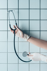 cropped shot of doctor in gloves holding stethoscope in front of tiled white wall