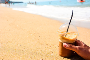 Ice coffee in a plastic glass on a yellow sand in a man's hand ready for a afternoon break and enjoying the beach.
