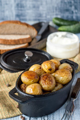 Baked young potatoes in a cast-iron pot, a jar of sour cream, fresh cucumbers and bread on an old wooden table. 