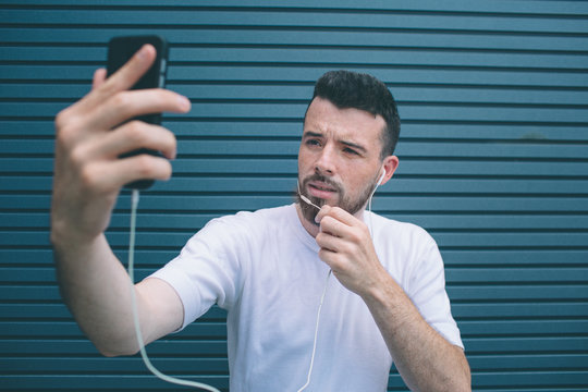 Guy is posing on camera. He is taking selfie and holding one hand on chin. Also man is listening to music. Isolated on striped and blue background.