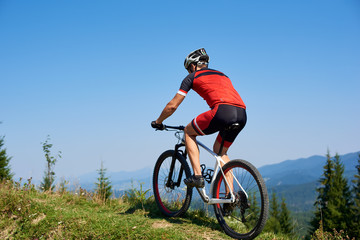 Fototapeta na wymiar Strong man tourist cyclist in helmet, sunglasses and full equipment riding bike on grassy hill. Mountains and blue summer sky on background. Active lifestyle and extreme sport concept