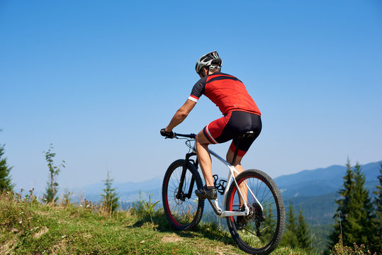 Strong man tourist cyclist in helmet, sunglasses and full equipment riding bike on grassy hill. Mountains and blue summer sky on background. Active lifestyle and extreme sport concept