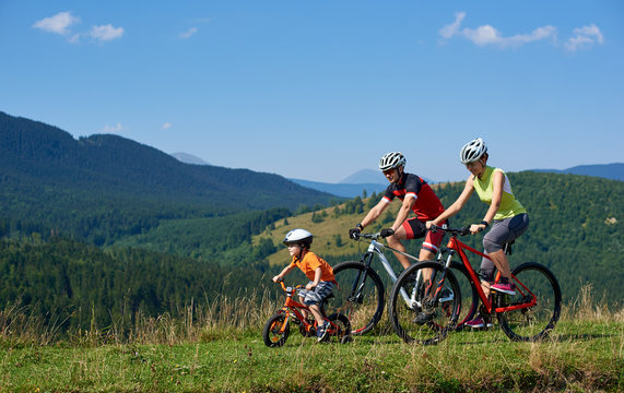 Sporty active family cyclists, mother, father and kid cycling on bikes on grassy hill. Carpathian mountains, blue summer sky on background. Healthy lifestyle, traveling and happy relations concept.