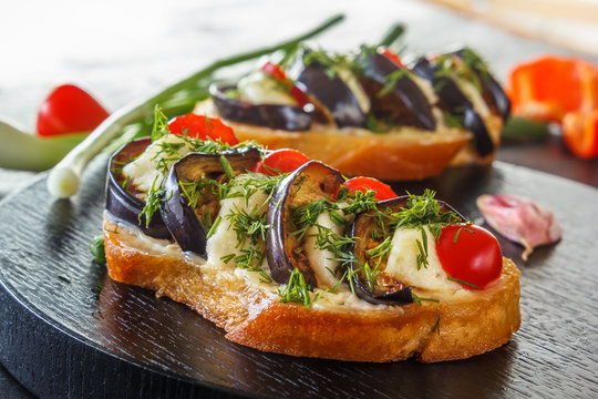 Bruschetta with fried eggplants, fresh tomatoes and cheese on black boards against the background of fresh vegetables. Close-up