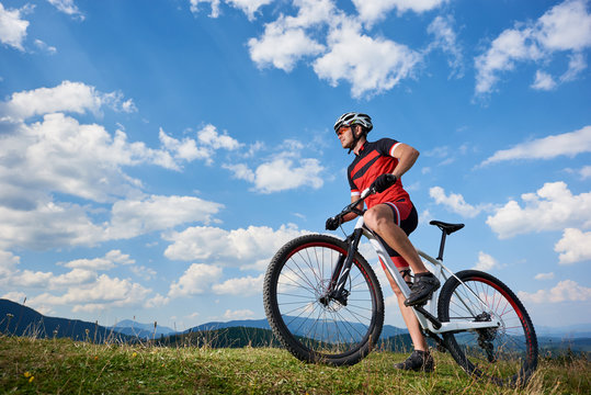 Profile of young athletic tourist biker in professional sportswear starting to cycle a bike on distant mountains and blue summer sky background. Active lifestyle and extreme sport concept.