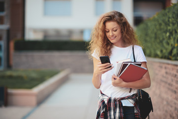 Female student using smart phone and holding books while walking in campus.