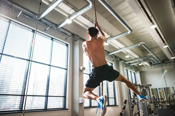 Fit young topless man in gym climbing a rope.