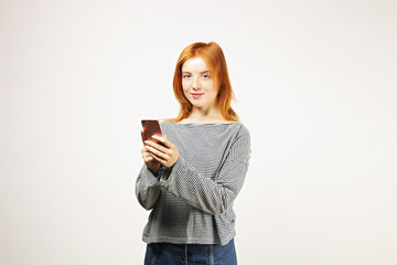 Fototapeta premium Young attractive natural redhead woman smiling at smartphone screen, using mobile phone, wearing loose gray shirt. White background, copy space. Beautiful female using online dating app concept.