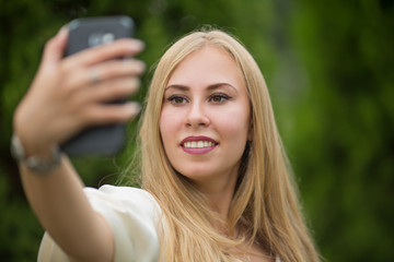 Young pretty blonde girl in a dress makes a selfie between thuja on the street in summer.