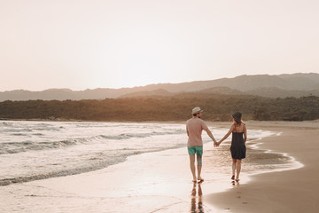 Back view of a romantic hipster couple walking at beach during summer vacation at sunset