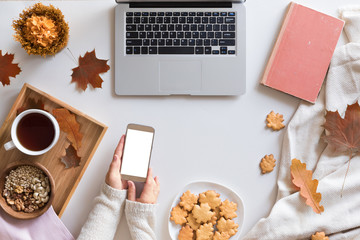 View from above of autumn background with mobile phone, laptop, autumn leaves, cookies, vintage book on white workspace. Top view and flat lay