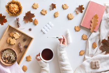 Naklejka na ściany i meble Top view and flat lay of autumn background with cold concept. Woman hands holding thermometer and pills, cup of hot tea, book, autumn leaves and flowers, cinnamon cookies on white background.