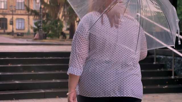 Fat ginger girl with glasses is alking up little stairs in park in rainy weather, holding umbrella