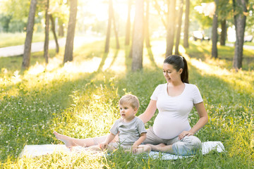 Young beautiful pregnant mother with baby son exercising and doing yoga on blue mat at summer park at sunset. Sportive and healthy motherhood. Fitness, happy maternity and healthy lifestyle concept.