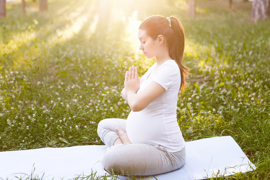 Pregnant woman doing yoga in lotus position in summer park on the green grass on fit mat, exercising and breathing outdoors. Healthy lifestyle and relaxation concept