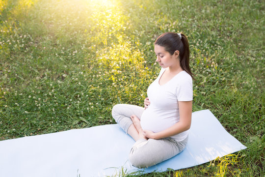 Young healthy pregnant woman doing yoga exercises in nature outdoors on green grass on fitness mat at sunset. Happy pregnancy and motherhood concept