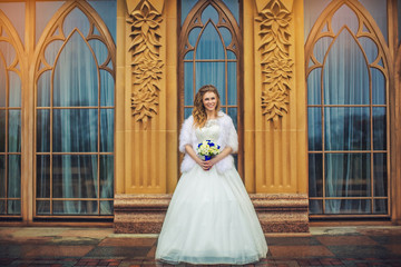 Obraz premium Happy bride holding wedding bouquet in hands on background of creative large windows, outdoors