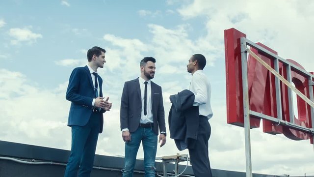 Three businessmen, two white and one african national, stend to edge of roof of skyscraper. Businessmen cheerfully discuss business. Share their achievements, remember funny stories. Top business.
