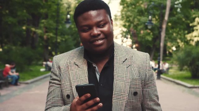 Joyful young urban professional Afro-American business man using smartphone while walking in city park. Having a date, feeling good, positive emotions. Joy of life, happiness. Male portrait, slow