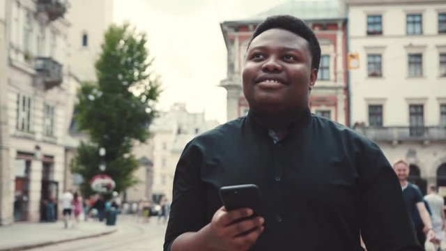 Cheerful young man in black formal shirt walks down the central city street, smiles, checks his phone, gets the message, reacts positively. Having fun, modern communication, devices, gadgets. Male