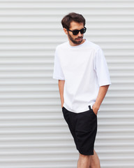Men's street youth modern fashion. A young male hipster in sunglasses with a beard posing against a white roller shutter blinds. White T-shirt and black shorts.