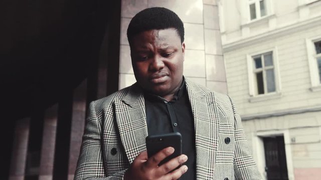 Young Afro-American businessman in a trendy jacket stands by the modern office, checks his phone, reacts negatively to received message, moves his head and actively gestures his hands in negotiation