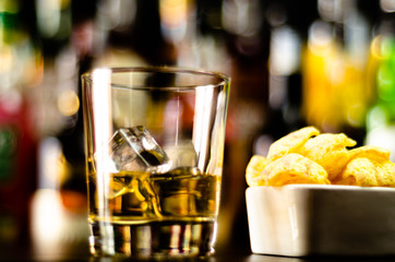 glass of whiskey with ice cubes and salty snacks on the background of the bar