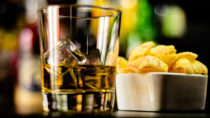 glass of whiskey with ice cubes and salty snacks on the background of the bar