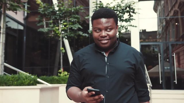 Cheerful young Afro-American man in black formal wear walks by the shopping center, smiling, actively using his smartphone. Positive emotions, good mood, enjoying the life. Happiness. Male portrait