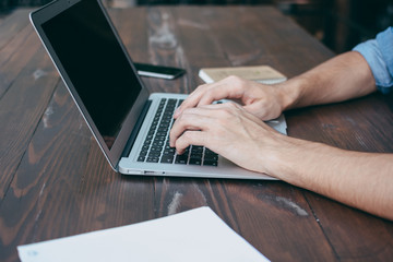 Men's hands are printed on a laptop on a wooden brown table in a cafe with free Wi-Fi, in the foreground a notebook. The concept for a freelancer works anytime, anywhere. Side view, concept.