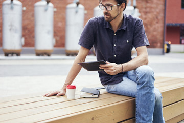 Young pretty guy in glasses working outside in park using modern tablet, notebook, mobile phone.