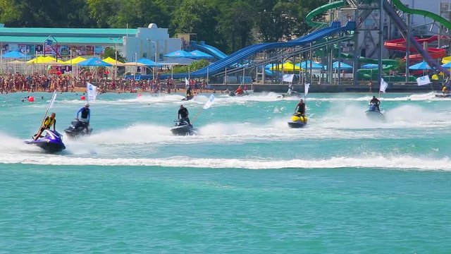 Hydrocycles with flags in row floats on azure surface of sea leaving trail of foam on background of sea shore with people