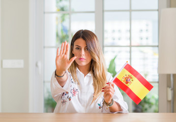 Young woman at home holding flag of Spain with open hand doing stop sign with serious and confident expression, defense gesture