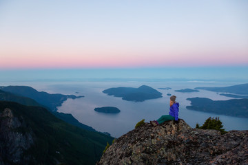 Fototapeta na wymiar Adventurous woman on top of a mountain cliff is enjoying the beautiful summer sunrise. Taken on Mount Brunswick, Lions Bay, North of Vancouver, BC, Canada.
