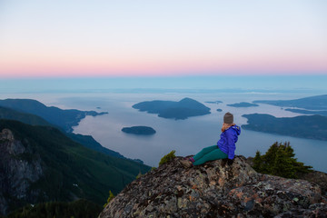 Naklejka premium Adventurous woman on top of a mountain cliff is enjoying the beautiful summer sunrise. Taken on Mount Brunswick, Lions Bay, North of Vancouver, BC, Canada.
