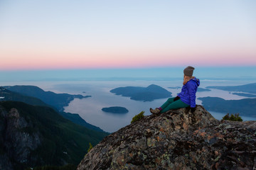 Adventurous woman on top of a mountain cliff is enjoying the beautiful summer sunrise. Taken on Mount Brunswick, Lions Bay, North of Vancouver, BC, Canada.