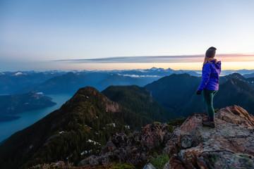 Fototapeta premium Adventurous woman on top of a mountain cliff is enjoying the beautiful summer sunrise. Taken on Mount Brunswick, Lions Bay, North of Vancouver, BC, Canada.