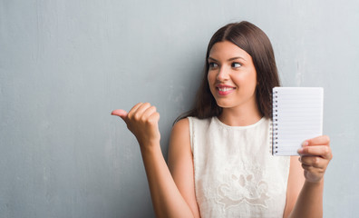Young brunette woman over grunge grey wall showing notebook pointing and showing with thumb up to...